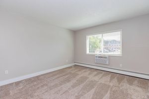 A light-colored room with beige carpet, featuring a single window with blinds. An air conditioning unit is mounted below the window. The walls are painted a soft gray, creating a neutral, spacious atmosphere. The room is empty, providing a blank canvas for furniture arrangement.