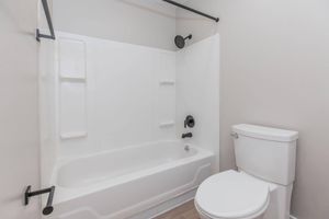 A clean, modern bathroom featuring a white bathtub with a shower head, a black curved shower rod, and a white toilet. The walls are light-colored, creating a spacious feel, and the floor appears to be a light wood finish.