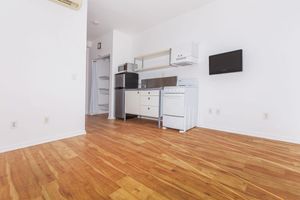 A modern, minimalist kitchen area featuring white walls, wooden flooring, and a compact setup. It includes a small refrigerator, a microwave, a stove, and a vent hood. A mounted TV is on the wall, and a doorway leads to a different room. The space is bright and uncluttered, emphasizing a clean design.