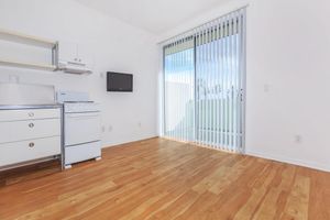 A bright, empty room with light wood flooring and white walls. It features a small kitchen area with a stove, a wall-mounted television, and large sliding glass doors with vertical blinds leading to an outdoor space. The room is spacious and well-lit.