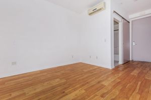 A spacious, empty room with light-colored walls and polished wooden flooring. An air conditioning unit is mounted on the wall, and a doorway leads to another space, partially visible. The overall ambiance is bright and minimalistic, suitable for various design options.