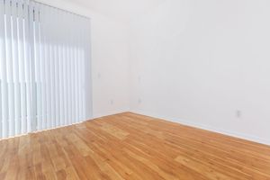 A bright, empty room with light-colored walls and wooden flooring. There are vertical blinds on a window letting in natural light, creating a spacious and airy atmosphere. The room is unfurnished, emphasizing the simplicity and potential for personalization.