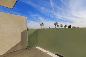 A view from a balcony featuring a green railing and a clear blue sky with wispy clouds. In the distance, several palm trees line the horizon, creating a peaceful and sunny atmosphere. The balcony surface is concrete, providing a modern and minimalistic aesthetic.