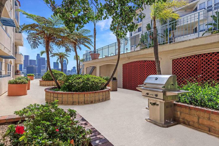A modern outdoor space featuring a stainless steel grill surrounded by lush greenery and flowing palm trees, with a view of a city skyline in the background.