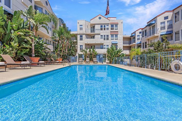 A bright blue swimming pool surrounded by lounge chairs and tropical plants, set in a well-maintained courtyard of an apartment community. The background features three-story buildings and a waving American flag under a clear blue sky. Ideal for relaxation and leisure activities.