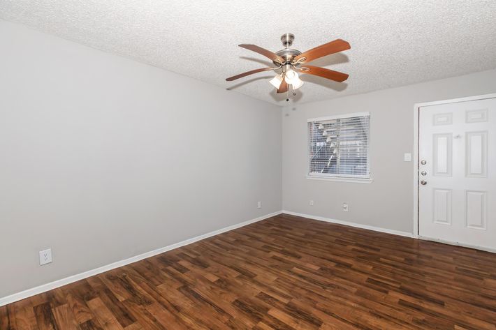 A vacant room with wooden flooring, a ceiling fan with wooden blades, a small window with blinds, and a white door. The walls are painted light gray, creating a bright and airy atmosphere.