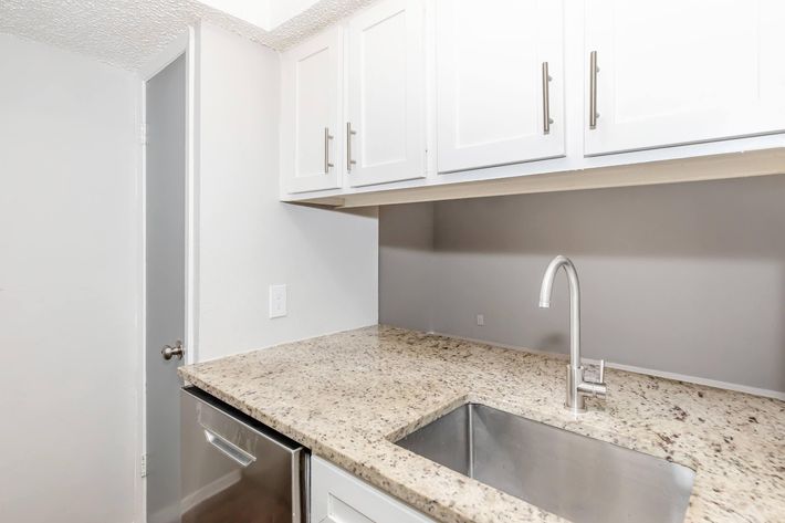 Interior view of a modern kitchen featuring a granite countertop, stainless steel sink, and white cabinets. The space is well-lit with light walls and a door visible in the background. The design showcases a clean and contemporary aesthetic, ideal for a functional cooking area.