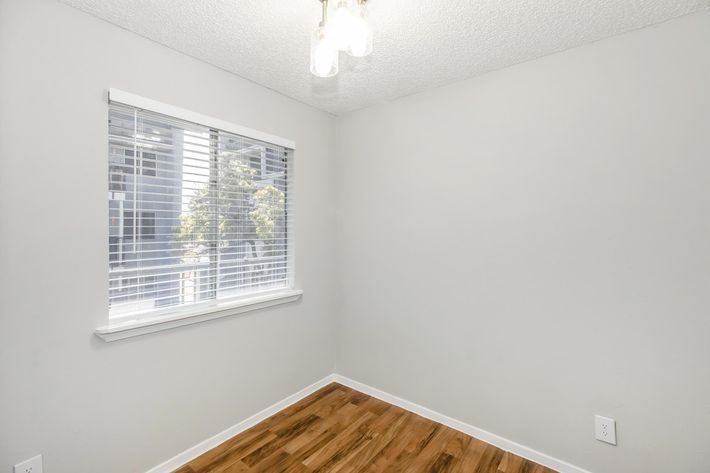 A small, empty room with light-colored walls and a wooden floor. A window with closed white blinds allows natural light to enter, and a simple light fixture hangs from the ceiling. The room appears clean and well-maintained, ready for decoration or furniture.