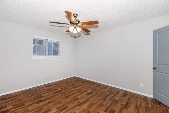 A well-lit, empty room with a fan featuring wooden blades on the ceiling. The walls are painted light gray, and there is a window with white blinds on one side. The floor is covered with modern wooden laminate flooring, and there's a gray door visible in the corner.