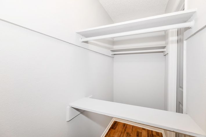 Empty closet with white walls and shelves, featuring a wooden floor. The space is well-lit and organized, with two horizontal shelves and an unobstructed view towards the back.