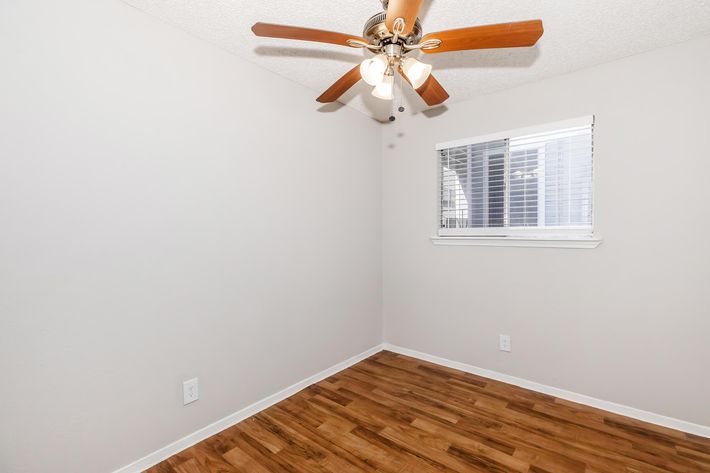 A simple, empty room featuring a ceiling fan with wooden blades, light fixtures, and a window with blinds. The walls are painted light gray, and the floor is covered with wooden laminate. The room is well-lit and uncluttered, providing a neutral and open space.