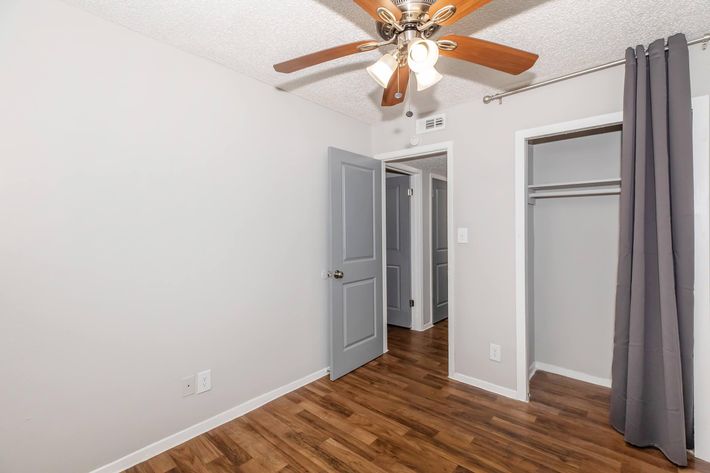 A well-lit room featuring a ceiling fan with wooden blades, light-colored walls, and hardwood flooring. An open door reveals a hallway, while another door leads to a closet with a rod. A gray curtain hangs beside the closet entrance, adding a touch of decor to the space.