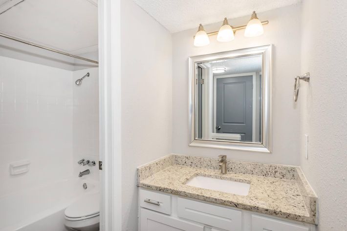 A modern bathroom featuring a white bathtub and shower combination, a sleek countertop with a rectangular sink, and a large mirror framed in silver. The lighting is provided by three light fixtures above the mirror, and there is a door leading to another room in the background.
