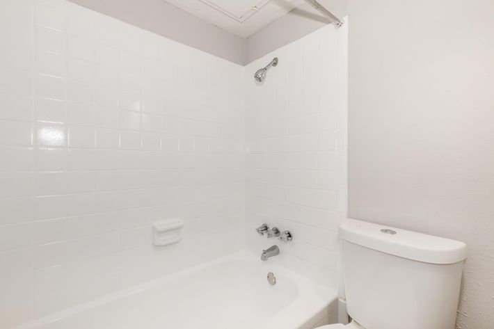 A clean, well-lit bathroom featuring a white bathtub with a showerhead, tiled walls, and a toilet. The space is simple and modern, with a neutral color palette emphasizing the bright, fresh appearance.