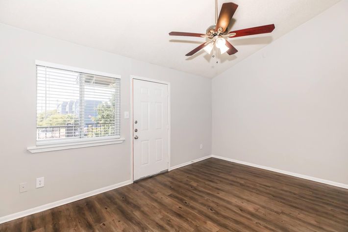 A well-lit room with a ceiling fan, featuring a light gray wall and wooden floor. A window allows natural light to enter, and there's a front door on one side. The space appears clean and empty, suitable for various uses.
