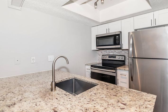 Modern kitchen featuring a granite countertop with an undermount sink, stainless steel appliances including a microwave, oven, and refrigerator, white cabinetry, and a stylish backsplash. The space is well-lit with overhead lighting and has a neutral color scheme.