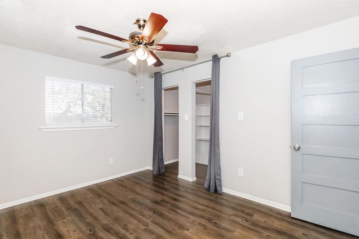 A clean, minimally decorated room featuring a ceiling fan, hardwood-like flooring, and light gray walls. A window with blinds allows natural light, while a closet is visible on one side. Gray curtains hang adjacent to the closet, and a light gray door is seen on the right.