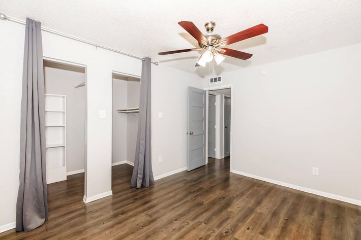 A well-lit interior of a bedroom featuring a ceiling fan, light gray walls, and dark wood laminate flooring. Two entrances are visible: one leads to a closet with shelves, and the other to a hallway. Light gray curtains hang on either side of the closet area.