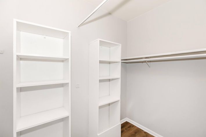 A spacious, empty closet featuring two white shelving units against a light gray wall. One shelving unit is located on the left side and the other on the right, with a hanging rod above for clothes storage. The floor is wooden, adding warmth to the minimalistic design.