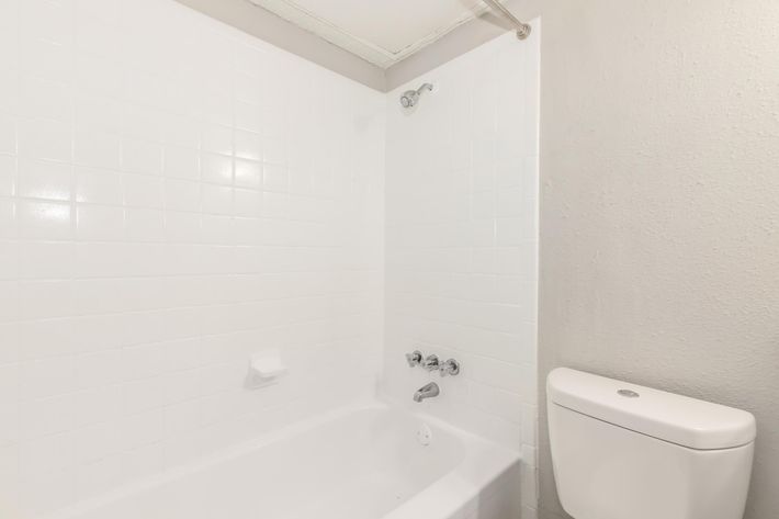 A clean, modern bathroom featuring a white tiled shower area and a bathtub. A chrome faucet and controls are visible on the wall. To the right, a white toilet is positioned next to the wall. The overall aesthetic is minimalist and bright, with neutral colors.