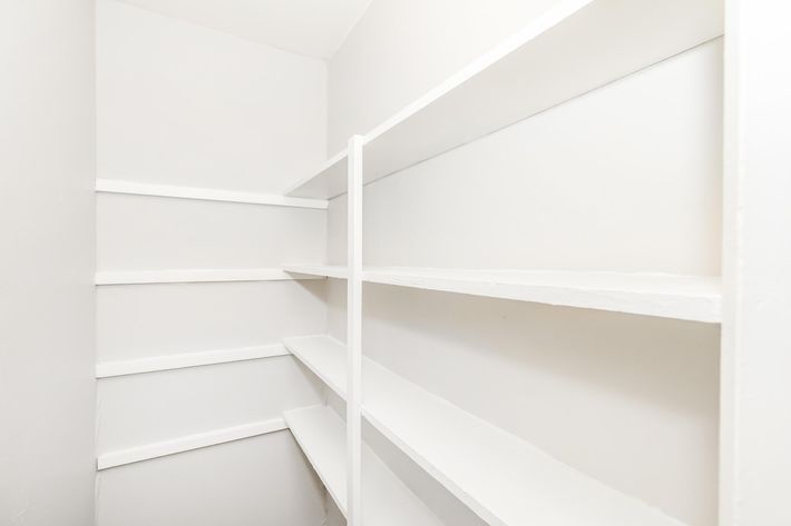A clean, empty pantry or storage closet with white walls and shelves. The shelves run along two sides, providing ample space for storage, but currently have no items on them. The overall appearance is minimalistic and organized.
