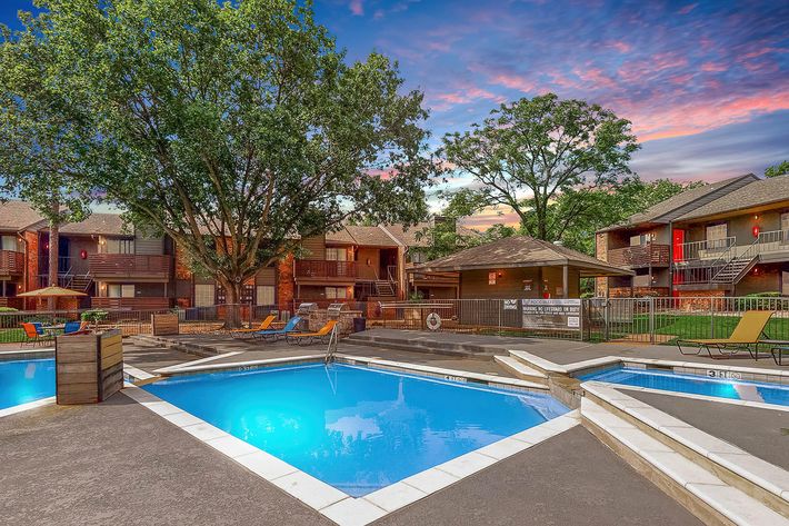 A well-maintained apartment community featuring a swimming pool surrounded by lounge chairs. The scene is enhanced by lush trees and a colorful sunset sky, with several residential buildings visible in the background. The pool area is inviting and well-lit, creating a relaxing atmosphere.