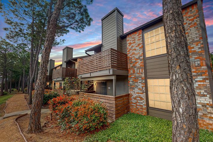 A view of a multi-unit residential building with brick and wood siding, featuring balconies and surrounded by lush greenery and colorful shrubs. The sky is painted with warm hues of sunset, adding a tranquil ambiance to the scene.