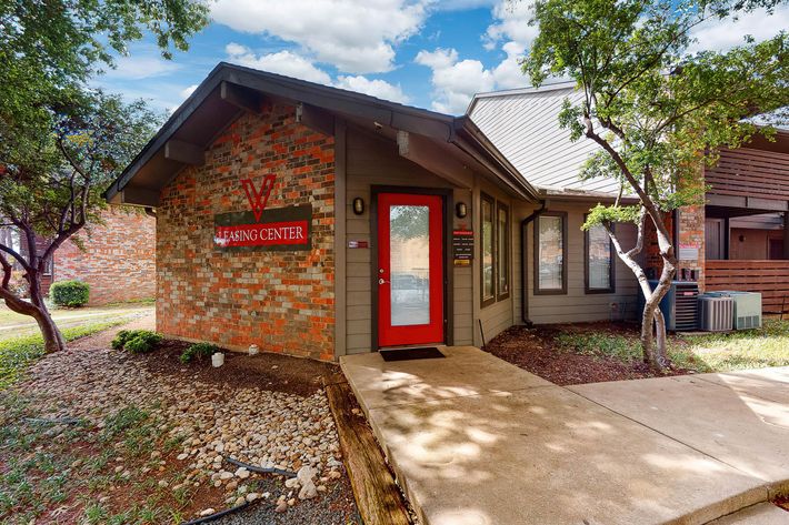 A small, one-story building with a brick façade and a red door labeled "Leasing Center." Surrounding the building is a landscaped area with trees and a stone pathway. In the background, there are other buildings and a blue sky with scattered clouds.