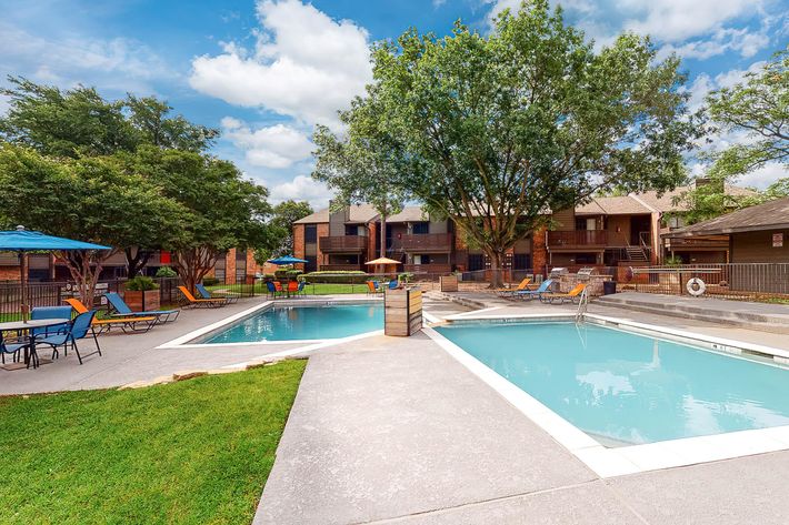 A well-maintained outdoor pool area featuring two swimming pools surrounded by lounge chairs and umbrellas. Lush green trees provide shade, and nearby, several apartment buildings can be seen. The sky is partly cloudy, creating a bright and inviting atmosphere.