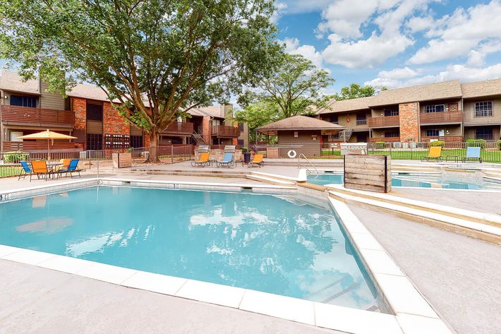 A swimming pool area surrounded by lounge chairs and shaded by trees, with two swimming pools separated by steps. In the background, there are apartment buildings with outdoor seating. The sky is blue with scattered clouds, creating a bright and inviting atmosphere.
