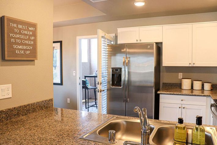 A modern kitchen with granite countertops and a stainless steel refrigerator. There are two bottles of olive oil on the counter near a sink. A motivational quote is framed on the wall beside an open door leading to a small outdoor space with a view of chairs and a table.