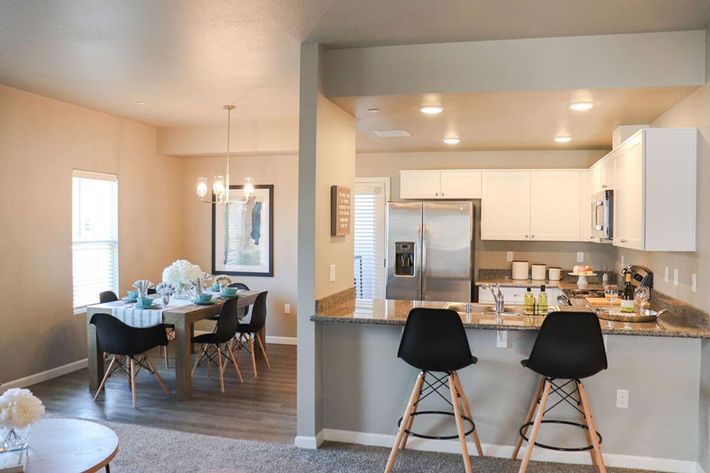 A modern open-concept kitchen and dining area featuring a granite countertop with bar stools, white cabinetry, stainless steel appliances, and a dining table set for a meal. Natural light floods the space through windows, highlighting a neutral color palette and stylish decor.