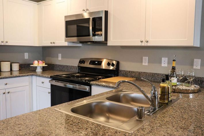 A modern kitchen featuring white cabinets, stainless steel appliances including a microwave and oven, a granite countertop, a dual sink, and a wooden cutting board. Decorative items like wine glasses and a bottle of olive oil are placed on the counter, adding a touch of elegance.