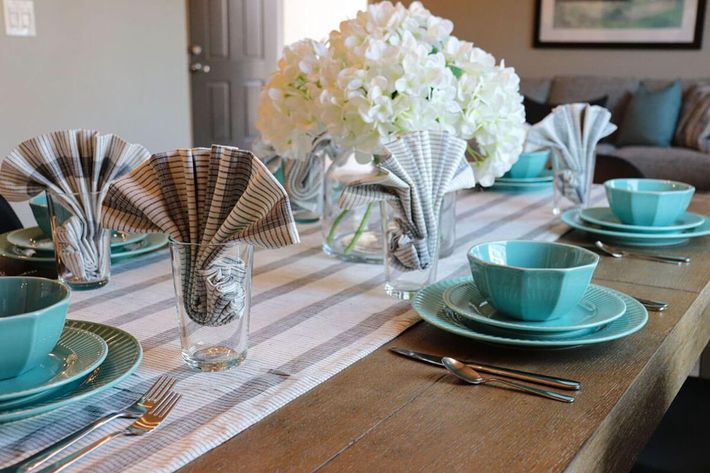 A beautifully set dining table featuring teal dishes, glassware, and neatly folded napkins. The table is adorned with a striped table runner and a centerpiece of white hydrangeas, creating an inviting and elegant ambiance. In the background, a cozy living area is visible.
