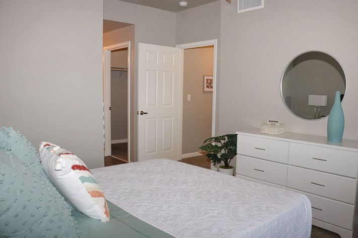 A well-lit bedroom featuring a bed with a decorative pillow, a white dresser, and a round mirror on the wall. A potted plant is positioned next to the dresser. There is a doorway leading to another room, and soft neutral-colored walls enhance the serene ambiance.