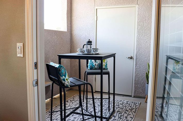 A modern small dining area featuring a tall black table with two high chairs. The table is set with a silver tray and a French press. Colorful patterned cushions are on the chairs, and the space is adorned with a light-colored rug. A door is visible in the background, along with a potted plant.