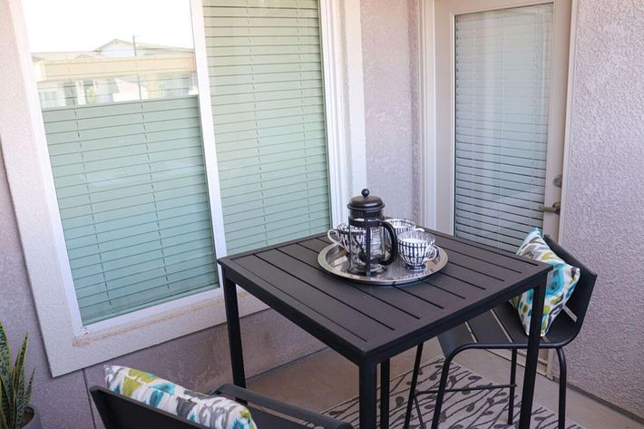 A small outdoor seating area featuring a square black metal table with a decorative tray holding a teapot and cups. Two chairs with patterned cushions are positioned around the table. The background includes large windows with blinds and a closed door, creating a cozy atmosphere.