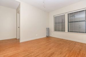 A spacious, empty room with light-colored walls and wooden flooring. There is a window with blinds on one wall, allowing natural light to enter. A door is visible on the left side, and a radiator is placed near the window. The overall ambiance is bright and uncluttered, suitable for various uses.