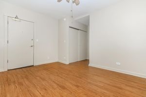 Empty room with light-colored walls and a wooden floor. A closed door is visible on the left, along with a closet door on the right. There is a ceiling fan overhead, but no furniture or decorations in the space, creating a minimalistic and open atmosphere.