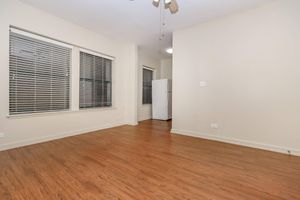 A spacious, empty room with light-colored walls and hardwood flooring. Large windows with slatted blinds allow natural light in. In the background, there is a white refrigerator against a wall. The room is well-lit and minimalistic, creating an inviting atmosphere.