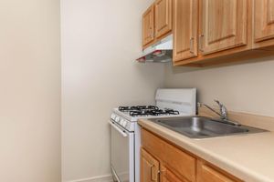 A small kitchen space featuring light-colored walls, wooden cabinets, a white gas stove with four burners, an exhaust hood, and a stainless steel sink with a single faucet, complemented by a beige countertop.