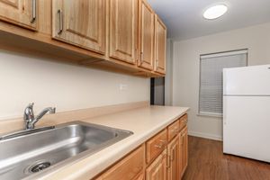 A clean kitchen with light wooden cabinets, a stainless steel sink, and a countertop. There's a white refrigerator beside a window with blinds. The floor is wooden, and overhead lighting provides a bright atmosphere.