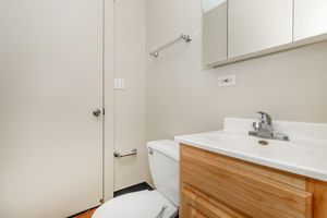 A clean, modern bathroom featuring a white toilet, a wooden cabinet under the sink, a simple faucet, and a wall-mounted mirror. The walls are painted light gray, and there is a towel rack and door visible. The floor has a dark finish, creating a contrast with the light-colored fixtures.