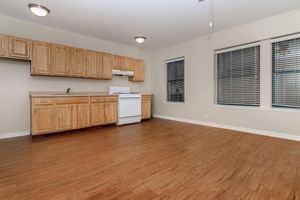 An empty kitchen and living area featuring wooden cabinets, a white refrigerator, and a stove. The floor is made of light hardwood, and there are large windows with blinds allowing natural light to fill the space. The walls are painted a neutral color, creating a spacious and inviting atmosphere.