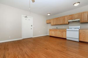 A spacious, empty kitchen area with light wood cabinets and a white stove. The floor is a warm, medium-toned hardwood. The room features a single door leading outside and overhead lighting, creating a bright and inviting atmosphere. The walls are painted in a soft beige color.