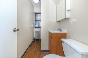 A clean, modern bathroom featuring a white toilet and a wooden vanity with a sink. There is a mirror above the sink, light-colored walls, and a window providing natural light. A radiator is positioned near the wall, and the floor is made of wooden planks.