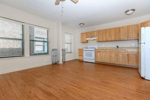 A spacious kitchen featuring wooden cabinets, a white stove, and a refrigerator. Natural light streams in through two large windows, illuminating the wood floor. A ceiling fan hangs above, and a vintage radiator stands against the wall, creating a warm and inviting atmosphere.