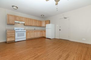 A spacious, well-lit kitchen featuring wooden cabinets, a white stove, and a refrigerator. The flooring is a warm, light wood, and there is an open area in front of the kitchen, allowing for easy movement and potential dining space. The walls are painted a soft neutral color.
