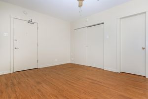 A light-colored empty room with wooden flooring, featuring a closed door on the left and a closet on the right. The walls are mostly bare, and there is a ceiling fan above. The space has a simple and minimal aesthetic, with natural light making the area feel open and inviting.