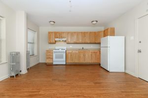 A modern, bright kitchen area featuring light wood cabinetry, a white stove, and a refrigerator. The flooring is a warm wood laminate, and there are large windows providing natural light. A vintage radiator is visible on the left, and the walls are painted in a neutral color, creating an inviting atmosphere.